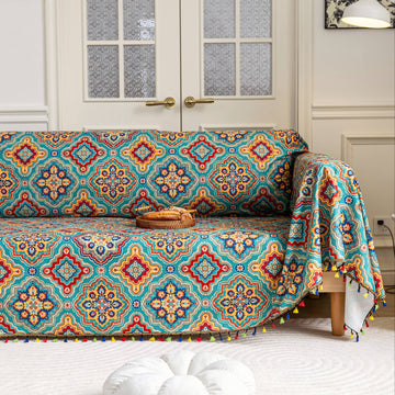 A vibrant, multicolored sofa cover featuring intricate geometric patterns in shades of red, blue, yellow, and green. The design includes detailed motifs and a playful fringe along the edges, adding a colorful and cozy touch to the living space.