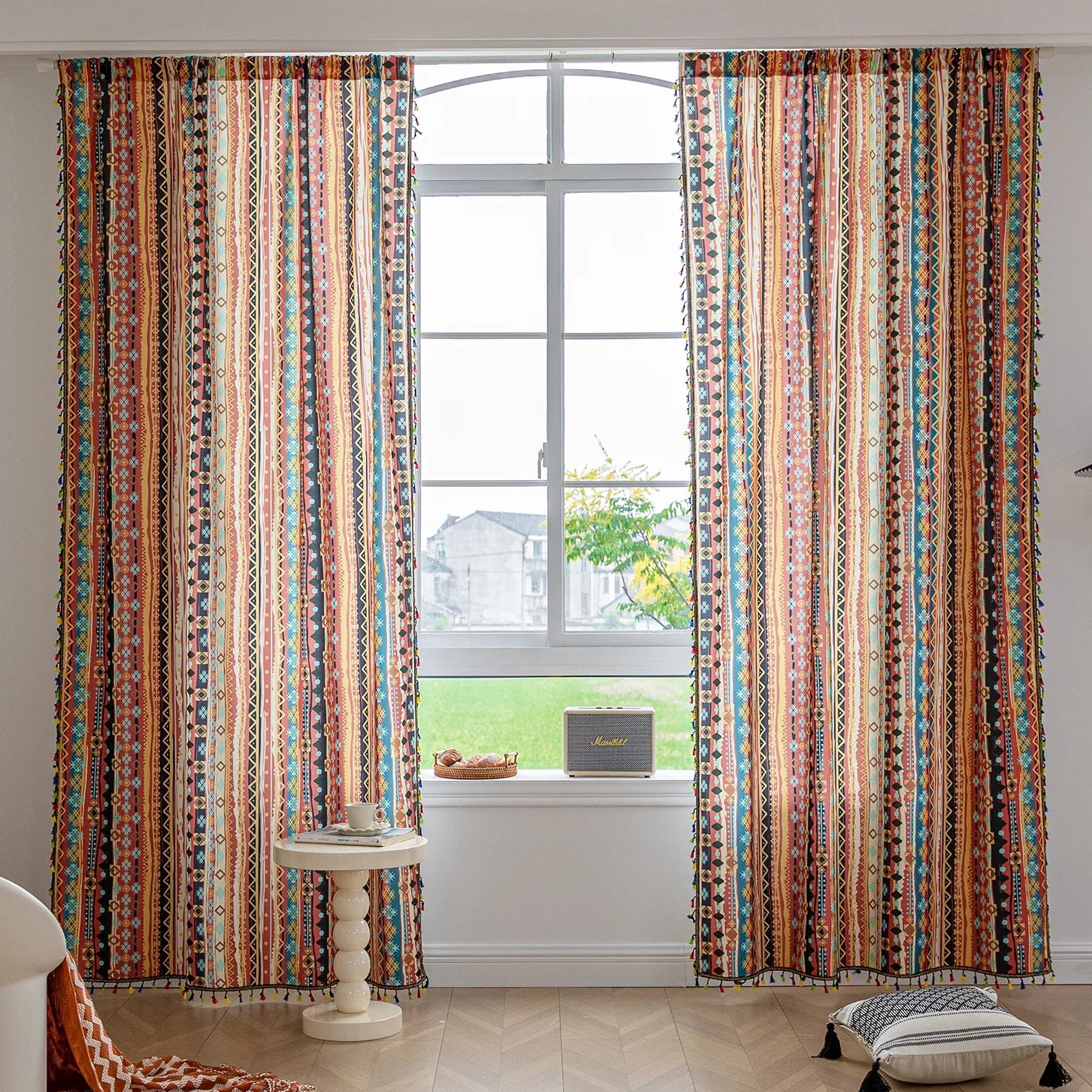 A vibrant pair of curtains featuring a bold geometric pattern in rich shades of red, orange, blue, and yellow. The design includes stripes with intricate shapes such as diamonds and circles, along with colorful tassels along the edges. The curtains frame a window with natural light streaming in, illuminating a cozy corner that includes a small side table with a cup and a portable speaker. A decorative cushion rests on the floor near the window.