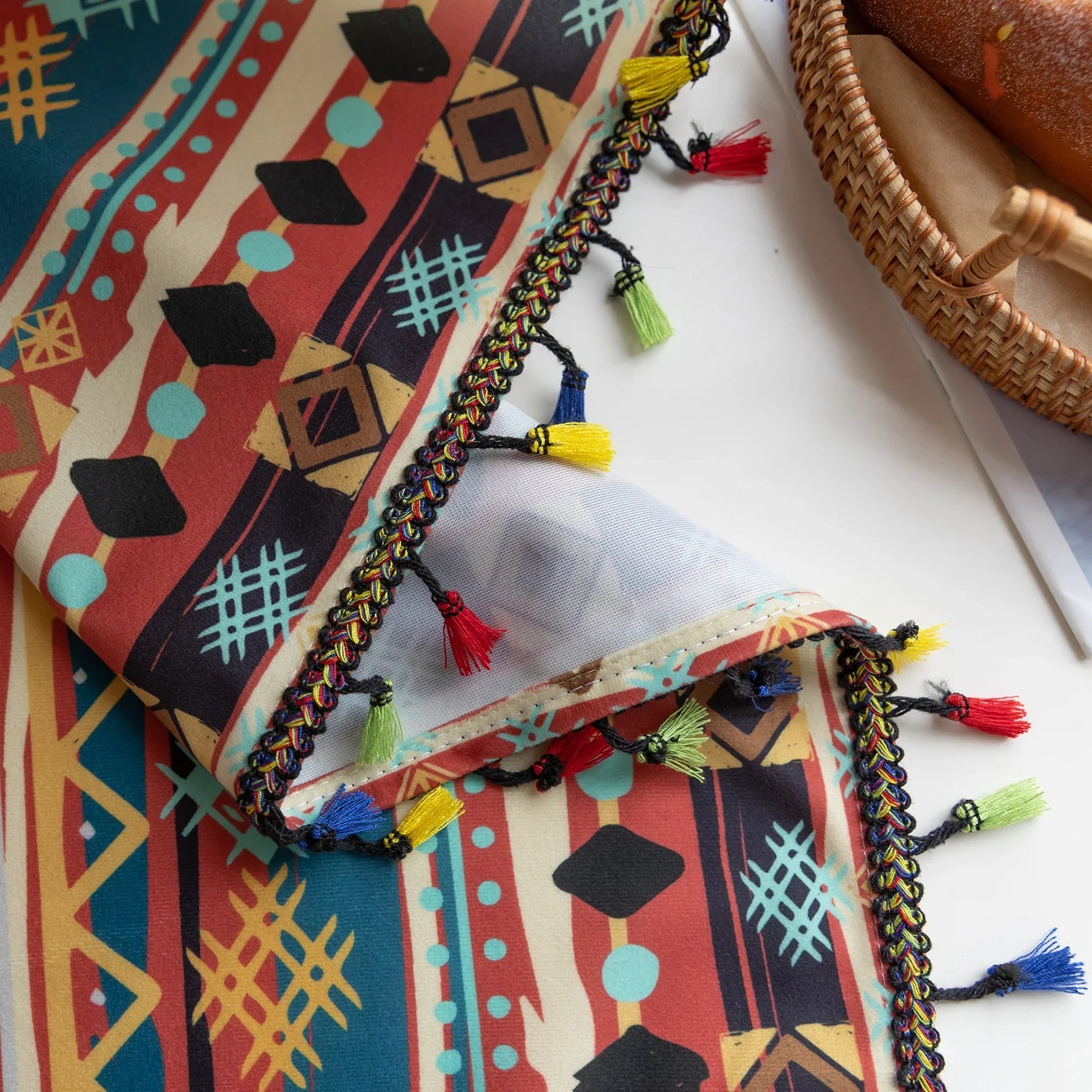 A close-up view of a colorful patterned fabric featuring bold geometric shapes in red, blue, and yellow. The fabric is edged with a decorative trim of colorful tassels in red, yellow, blue, and green, adding a playful and vibrant touch.