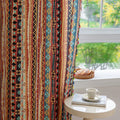 A colorful curtain with a vibrant geometric pattern featuring stripes of red, yellow, blue, and black. The design includes bold diamonds, circles, and lines, and is finished with playful tassels in multiple colors along the edges. The curtain is illuminated by natural light streaming through a window, creating a cozy and lively atmosphere in the room. A cup of tea and a stack of books rest on a side table near the window.