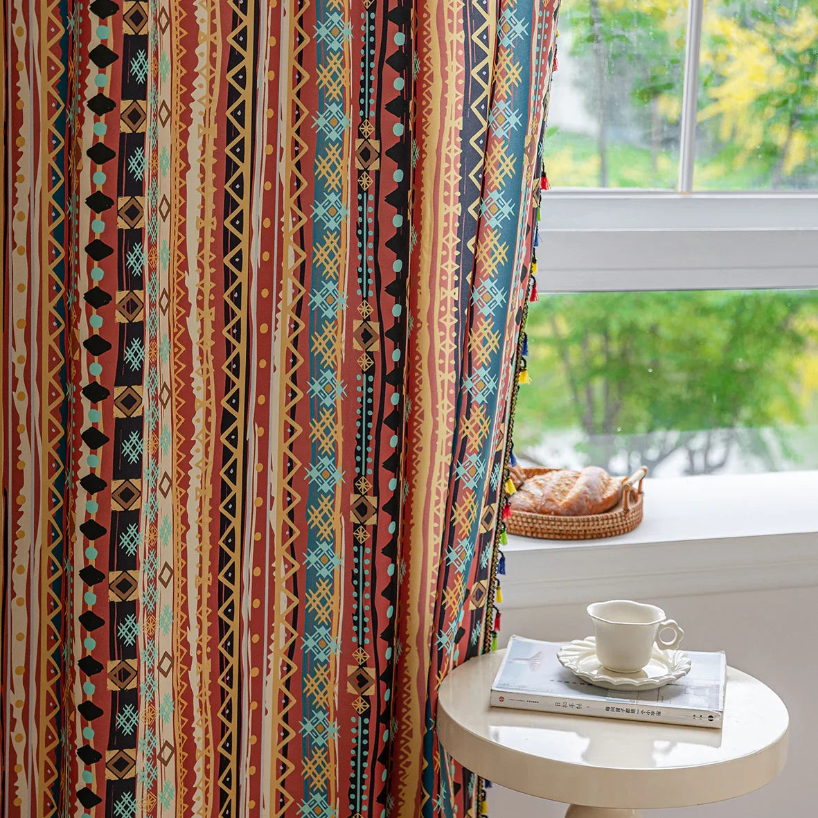 A colorful curtain with a vibrant geometric pattern featuring stripes of red, yellow, blue, and black. The design includes bold diamonds, circles, and lines, and is finished with playful tassels in multiple colors along the edges. The curtain is illuminated by natural light streaming through a window, creating a cozy and lively atmosphere in the room. A cup of tea and a stack of books rest on a side table near the window.