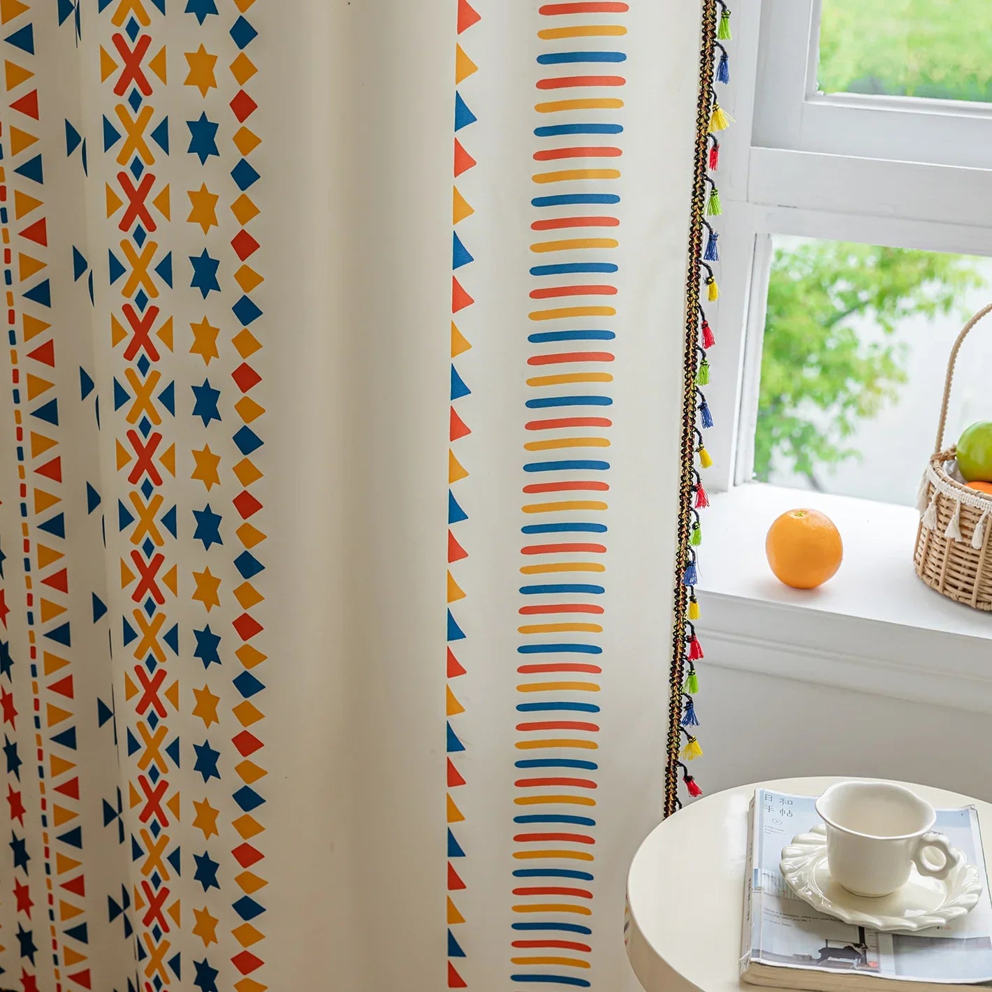 Colorful geometric patterned curtain with rows of blue, yellow, and red shapes beside a window, styled with a tasseled edge.