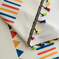 Close-up of white curtain fabric with colorful geometric prints in red, yellow, and blue, featuring a decorative black braided trim with small multicolor tassels for a playful, bohemian style.