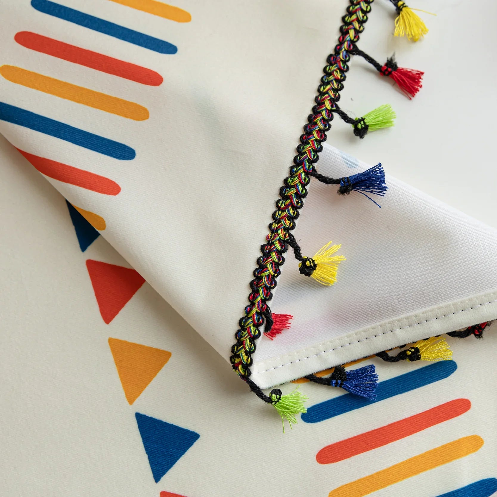 Close-up of white curtain fabric with colorful geometric prints in red, yellow, and blue, featuring a decorative black braided trim with small multicolor tassels for a playful, bohemian style.