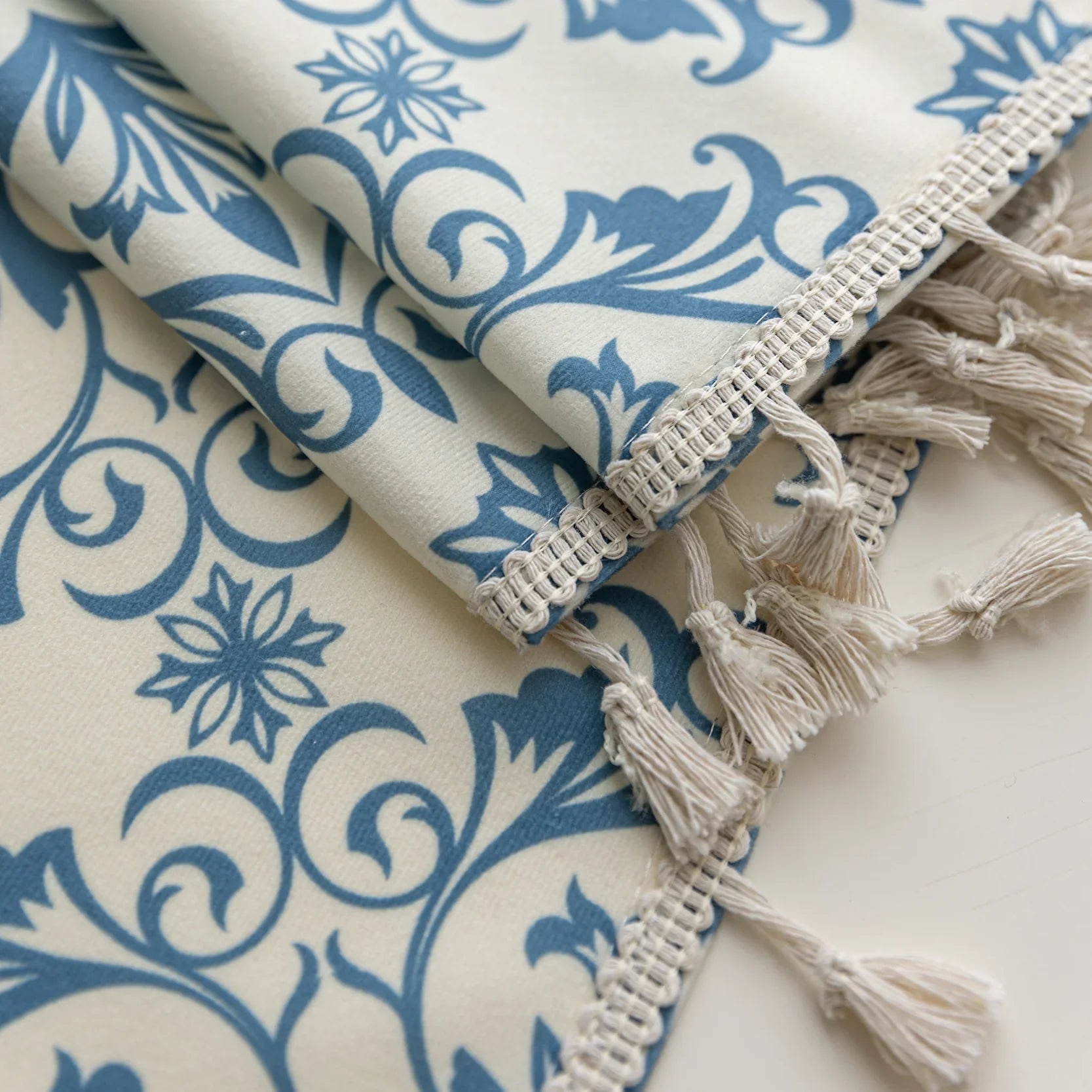Elegant cream curtain fabric with intricate blue floral patterns, finished with decorative beige tassel trim for a classic, sophisticated look.