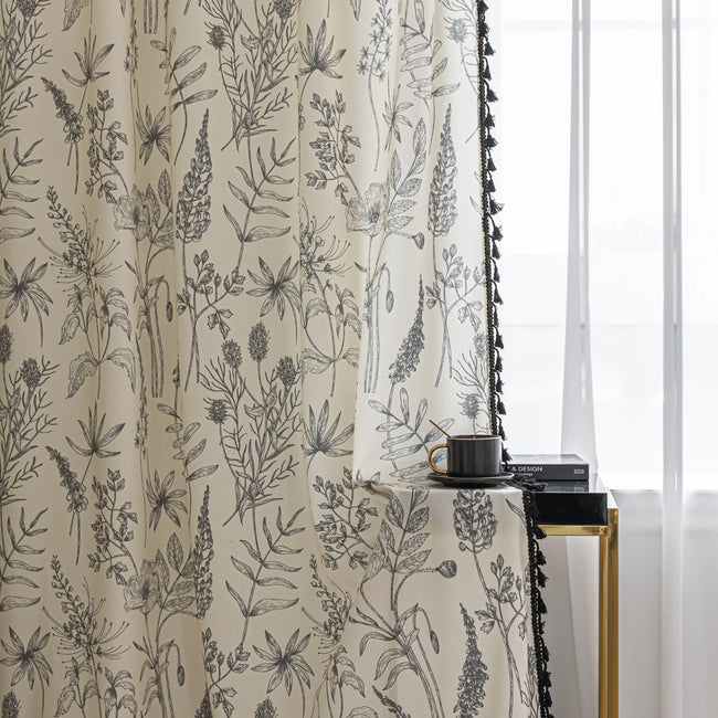 Cream curtain with black botanical print and black tassel trim, hanging beside a sheer white curtain.
