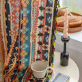 A vibrant pair of curtains featuring a bold geometric pattern in rich shades of red, orange, blue, and yellow. The design includes stripes with intricate shapes such as diamonds and circles, along with colorful tassels along the edges. The curtains frame a window with natural light streaming in, illuminating a cozy corner that includes a small side table with a cup and a portable speaker. A decorative cushion rests on the floor near the window.