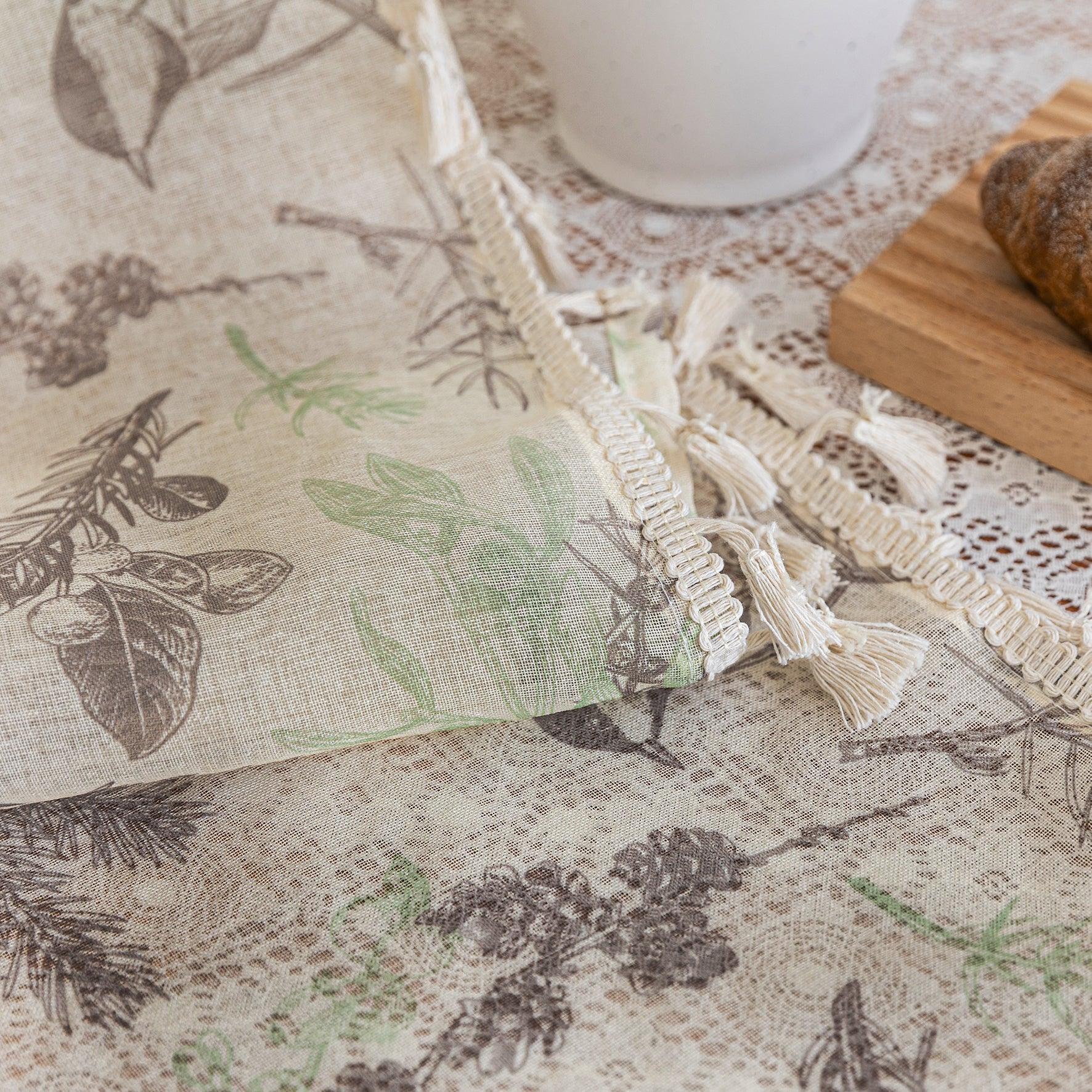 Close-up of beige linen curtain fabric with brown and green botanical and bird print, featuring decorative tassel trim, displayed on a lace tablecloth for a natural, rustic look.