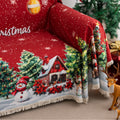 Festive red Christmas-themed sofa cover featuring decorated ornaments, snowflakes, snowy pine trees, a cheerful snowman, Santa Claus with gifts, and a cozy snow-covered house with poinsettias in the foreground.