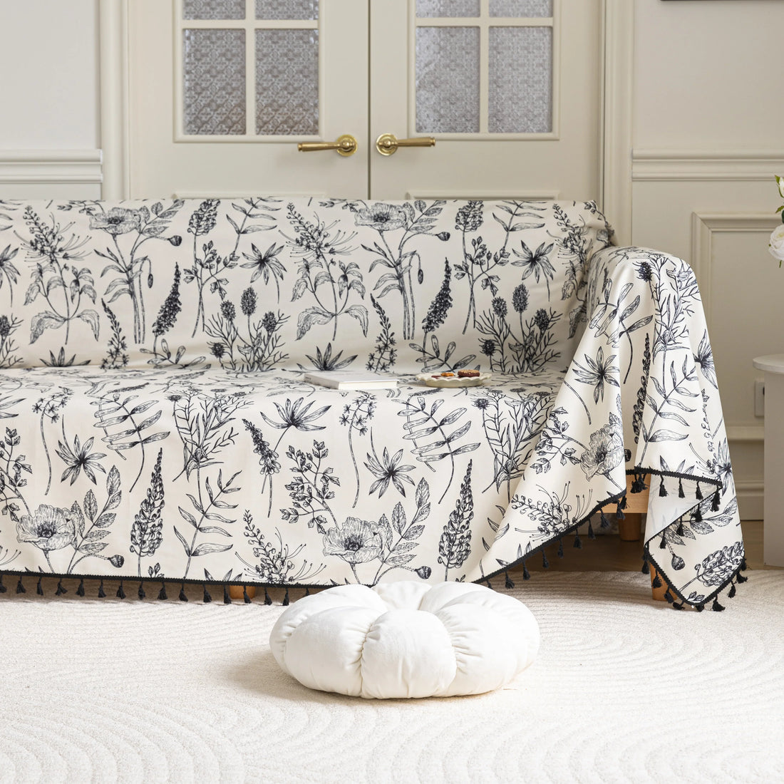 Elegant sofa cover with black botanical floral print on a cream background, featuring. delicate line-drawn flowers and plants with black tassel trim