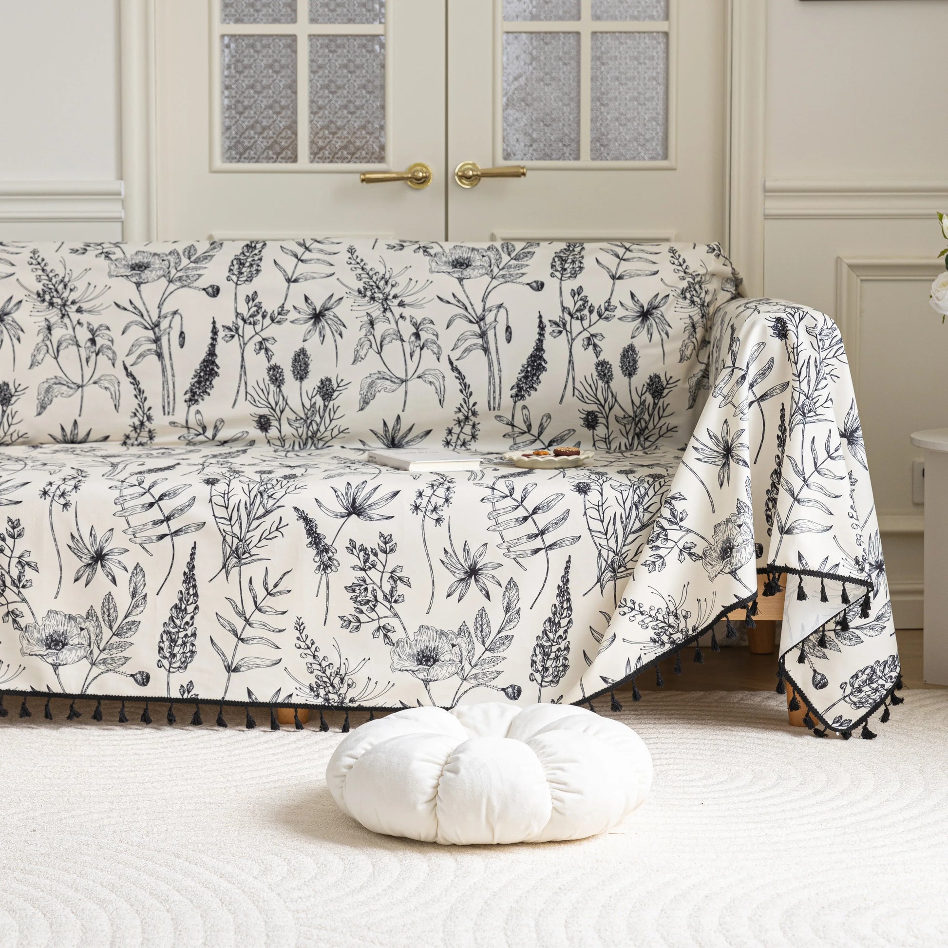 Elegant sofa cover with black botanical floral print on a cream background, featuring. delicate line-drawn flowers and plants with black tassel trim