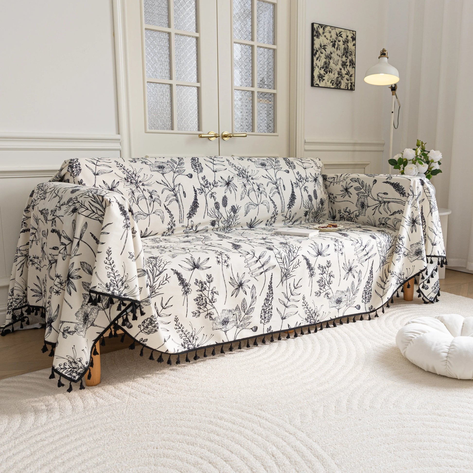 Elegant sofa cover with black botanical floral print on a cream background, featuring. delicate line-drawn flowers and plants with black tassel trim