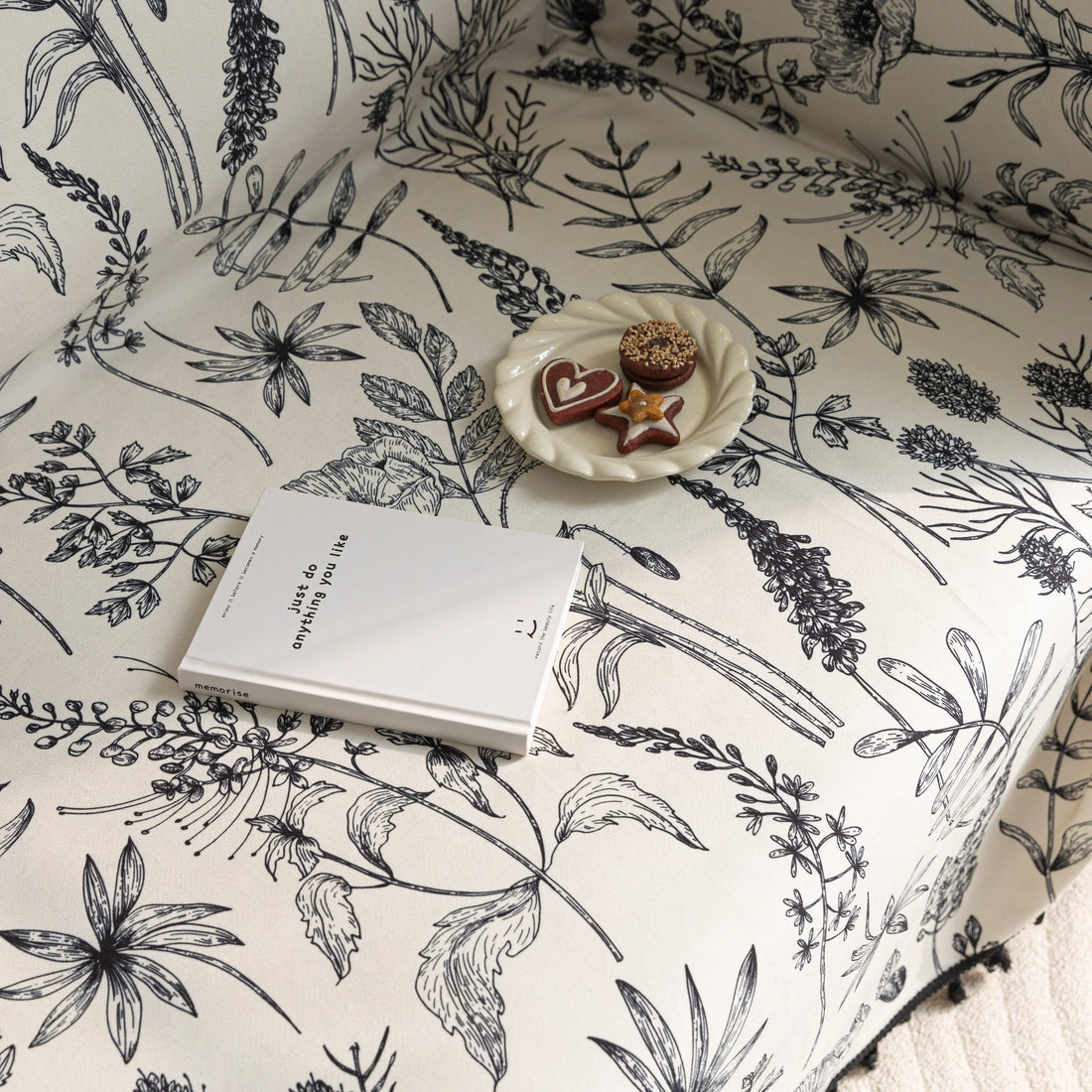 Elegant sofa cover with black botanical floral print on a cream background, featuring. delicate line-drawn flowers and plants with black tassel trim