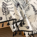 Elegant sofa cover with black botanical floral print on a cream background, featuring. delicate line-drawn flowers and plants with black tassel trim