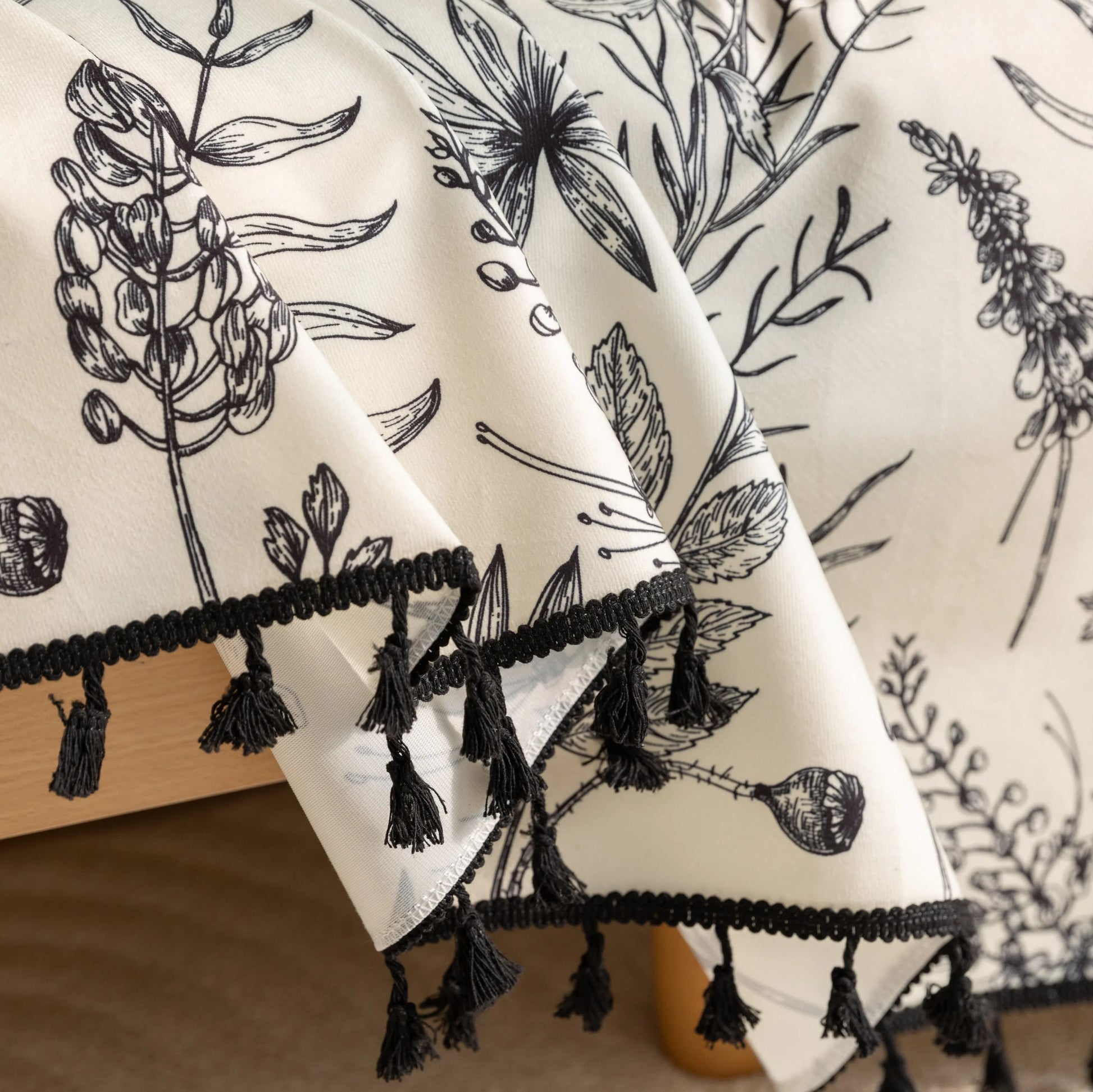 Elegant sofa cover with black botanical floral print on a cream background, featuring. delicate line-drawn flowers and plants with black tassel trim