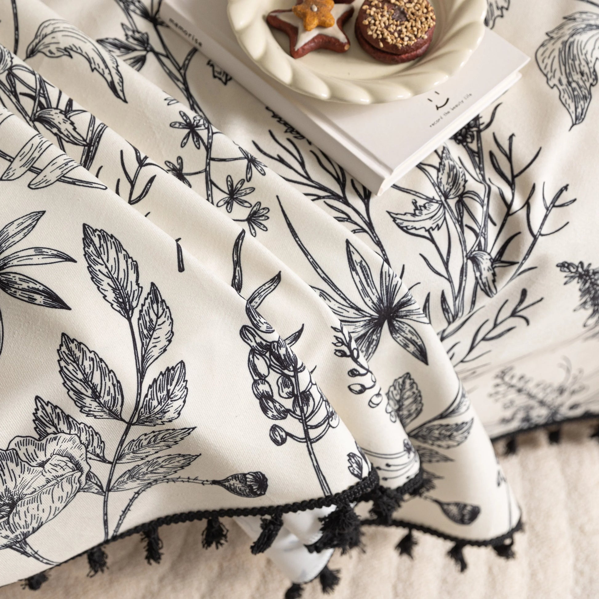 Elegant sofa cover with black botanical floral print on a cream background, featuring. delicate line-drawn flowers and plants with black tassel trim