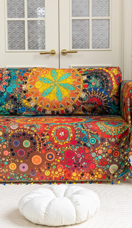 Colorful sofa cover with abstract circular and mandala-inspired patterns in vibrant shades of yellow, red, turquoise, and orange on a dark background, accented with multicolor tassel trim.