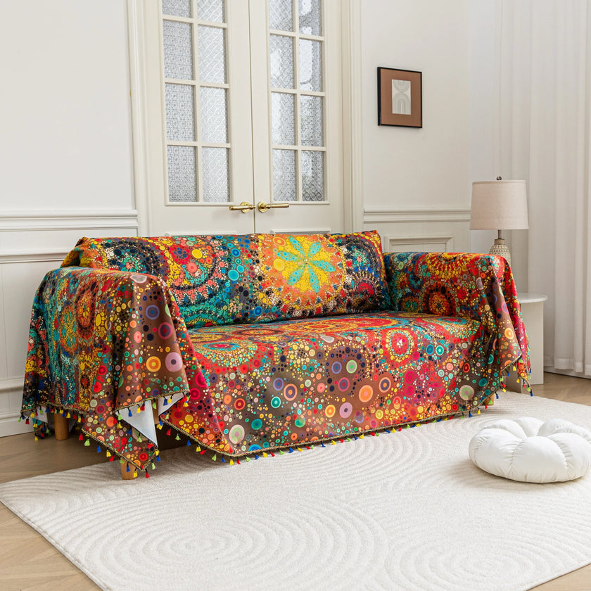 Colorful sofa cover with abstract circular and mandala-inspired patterns in vibrant shades of yellow, red, turquoise, and orange on a dark background, accented with multicolor tassel trim.