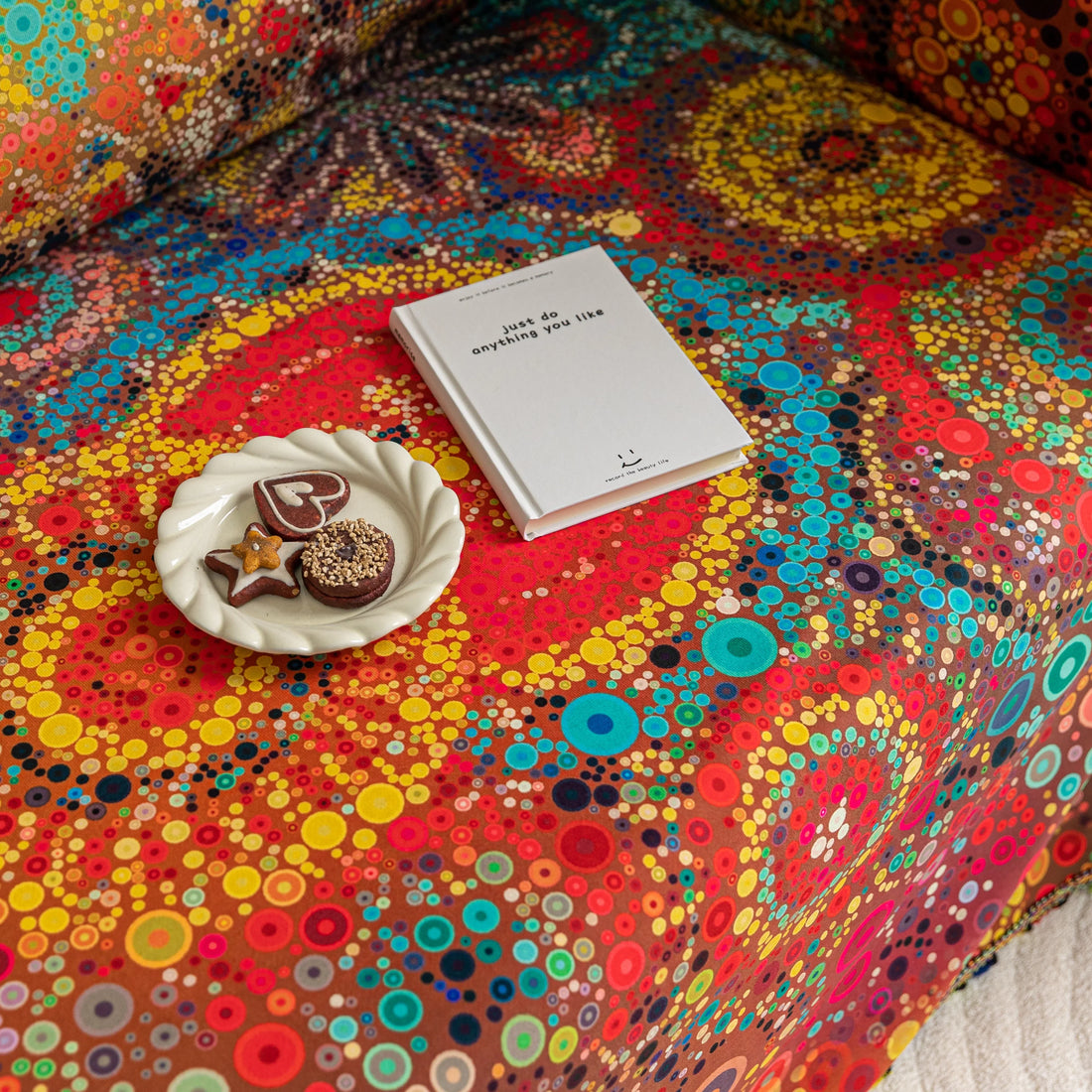 Colorful sofa cover with abstract circular and mandala-inspired patterns in vibrant shades of yellow, red, turquoise, and orange on a dark background, accented with multicolor tassel trim.