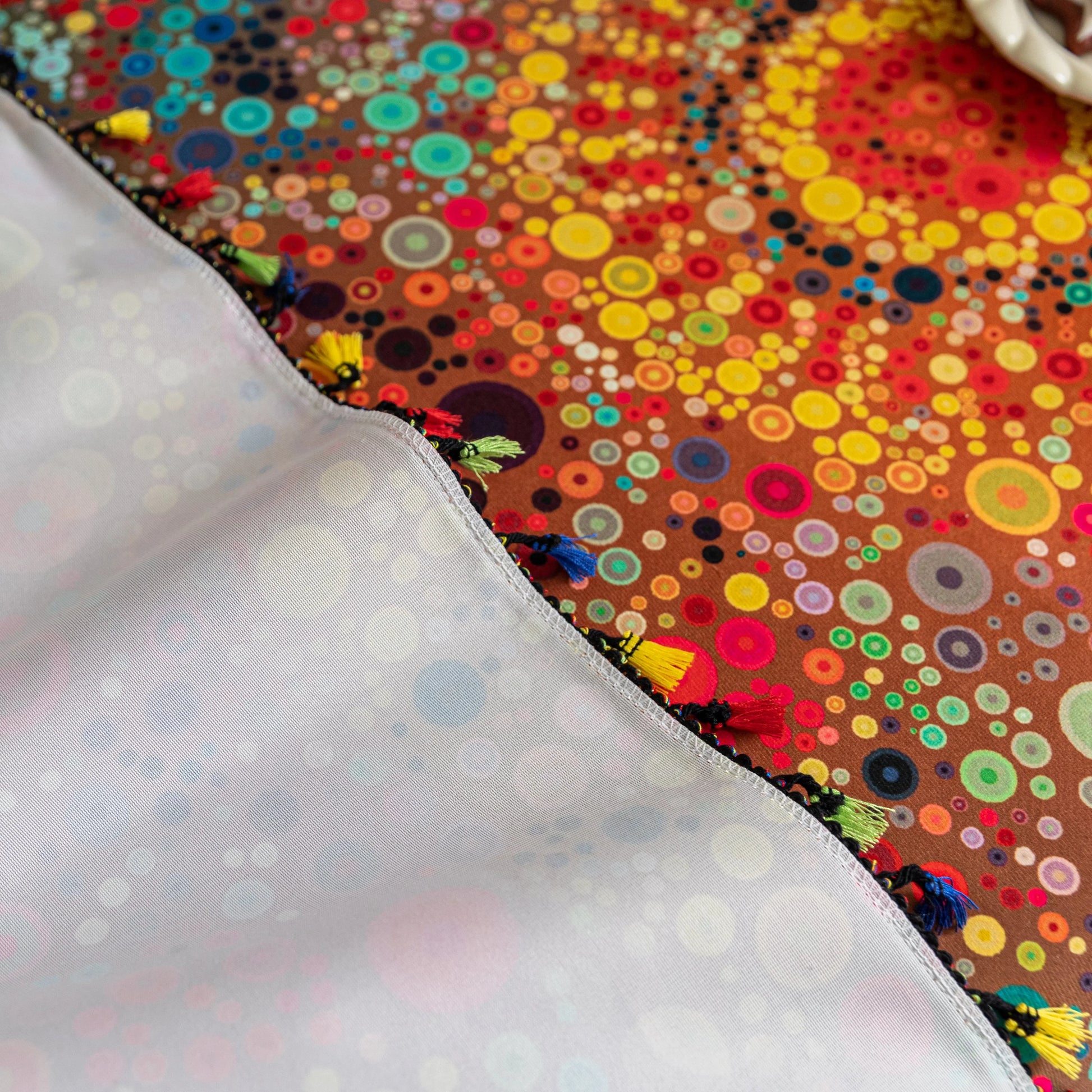 Colorful sofa cover with abstract circular and mandala-inspired patterns in vibrant shades of yellow, red, turquoise, and orange on a dark background, accented with multicolor tassel trim.