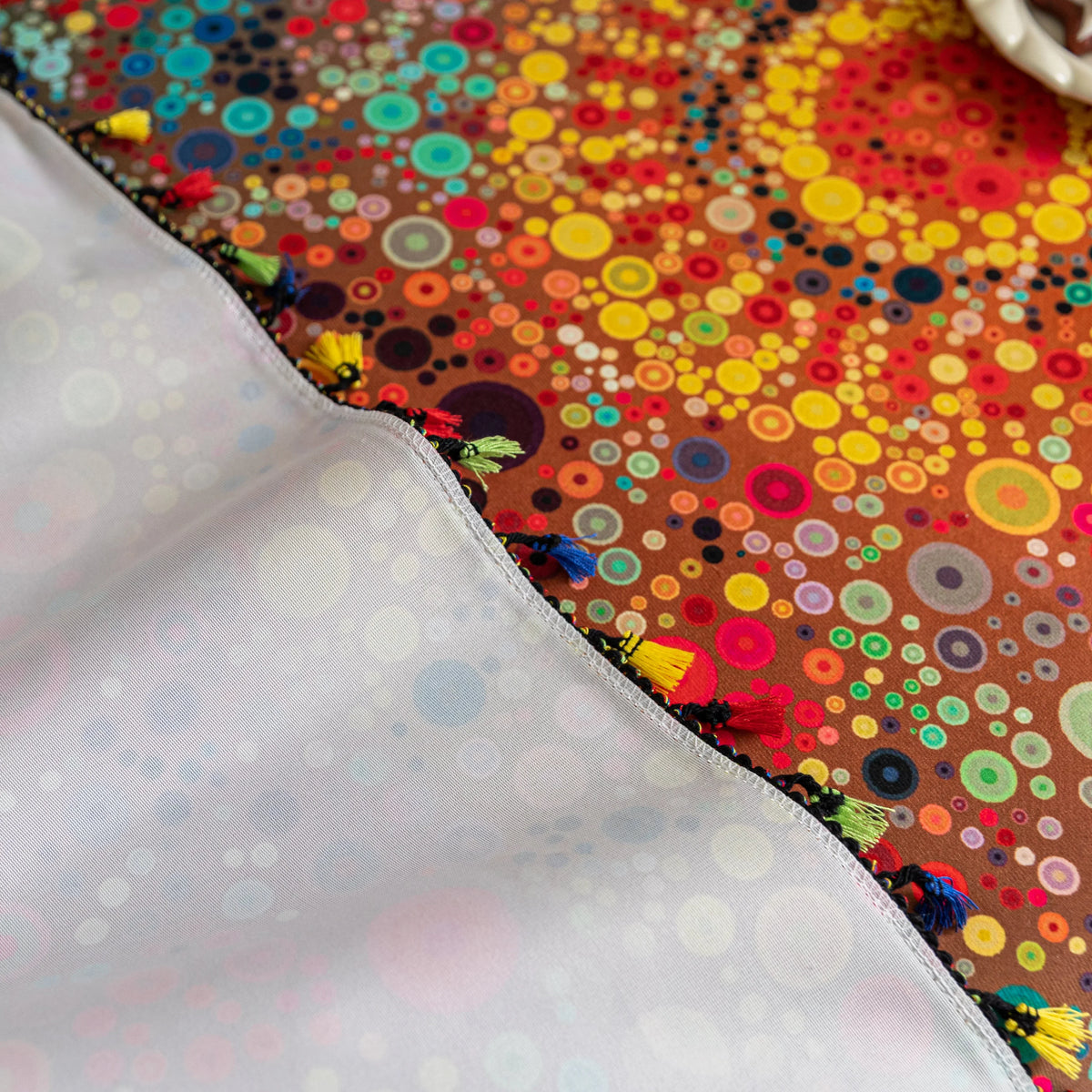 Colorful sofa cover with abstract circular and mandala-inspired patterns in vibrant shades of yellow, red, turquoise, and orange on a dark background, accented with multicolor tassel trim.