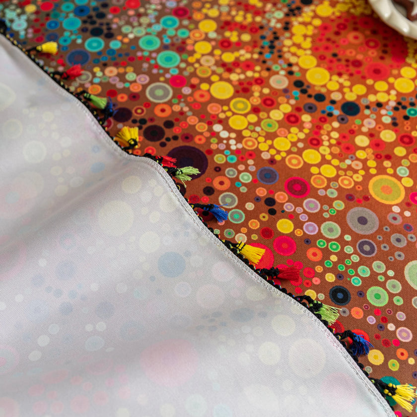 Colorful sofa cover with abstract circular and mandala-inspired patterns in vibrant shades of yellow, red, turquoise, and orange on a dark background, accented with multicolor tassel trim.