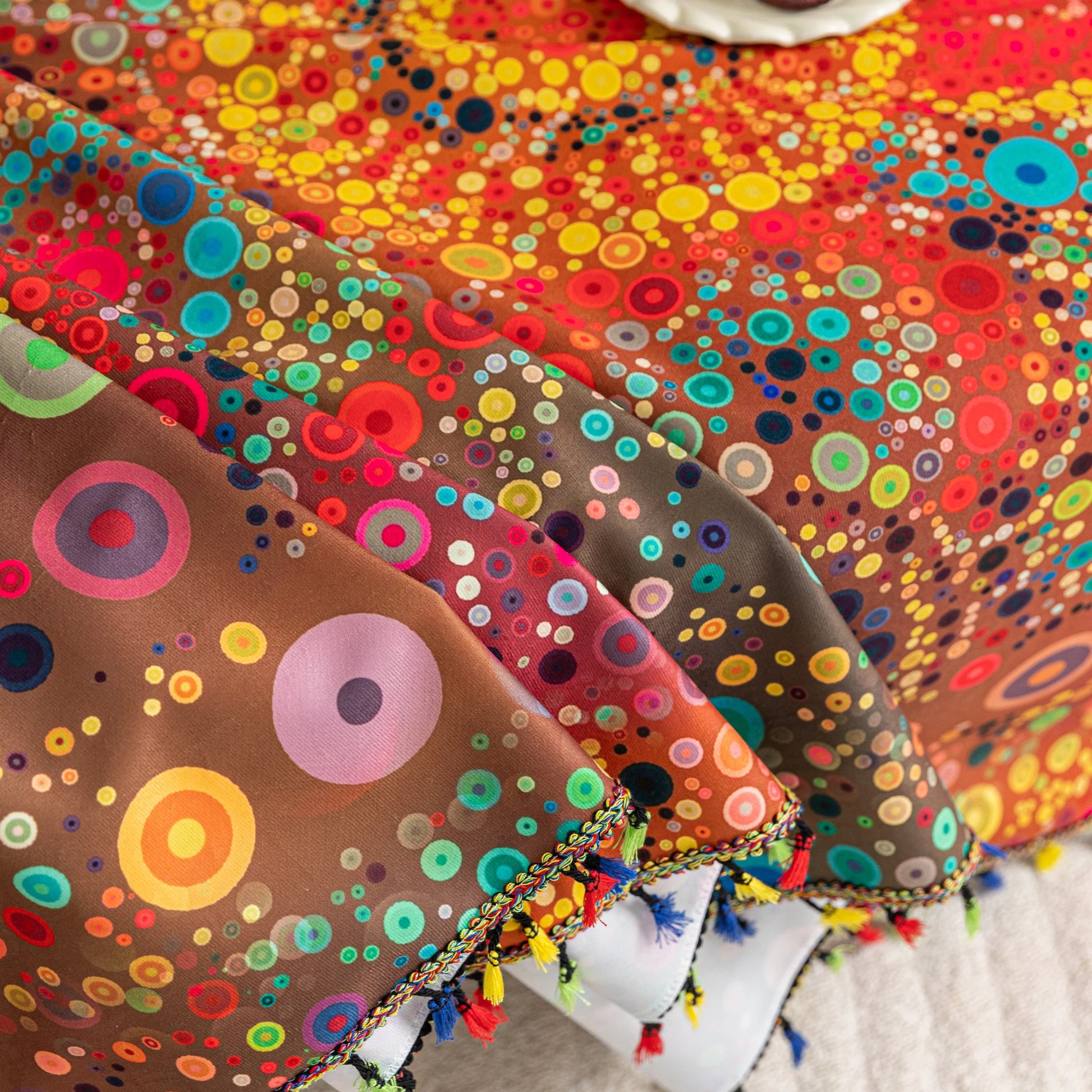 Colorful sofa cover with abstract circular and mandala-inspired patterns in vibrant shades of yellow, red, turquoise, and orange on a dark background, accented with multicolor tassel trim.