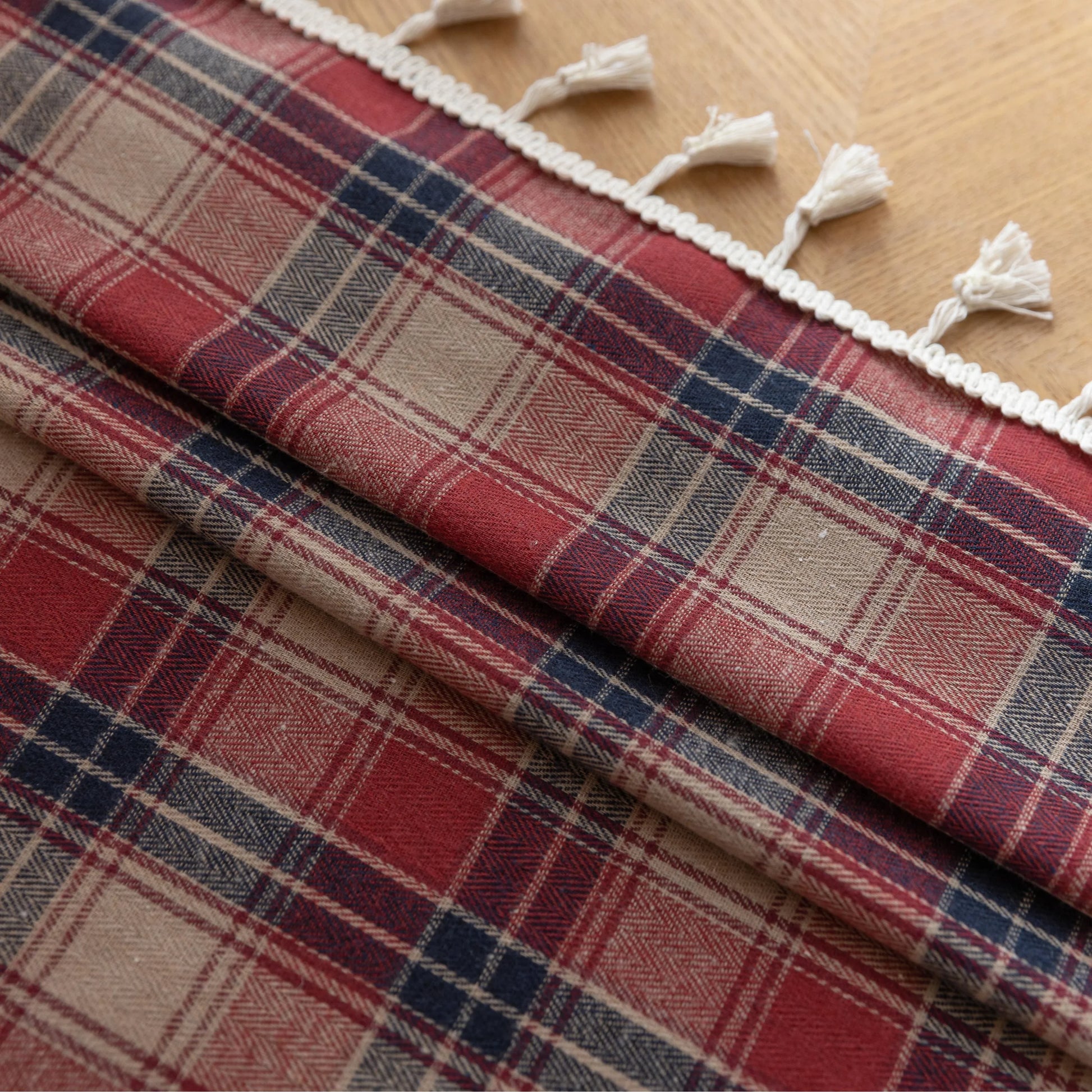 Red, navy blue, and beige plaid curtain fabric with a soft, textured weave and decorative white tassel trim on the edge.