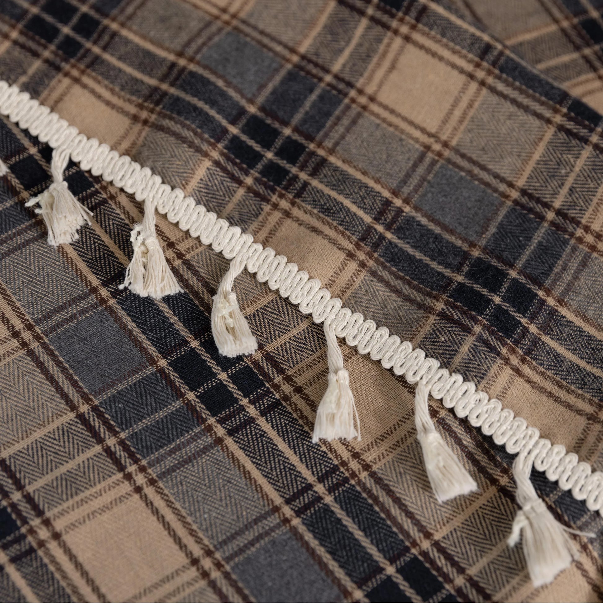 Beige and navy blue plaid curtain fabric with a soft, textured weave and decorative white tassel trim along the edge.