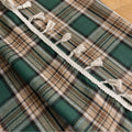 Green and beige plaid curtain fabric with a soft, woven texture, accented by decorative white tassel trim along the edge.