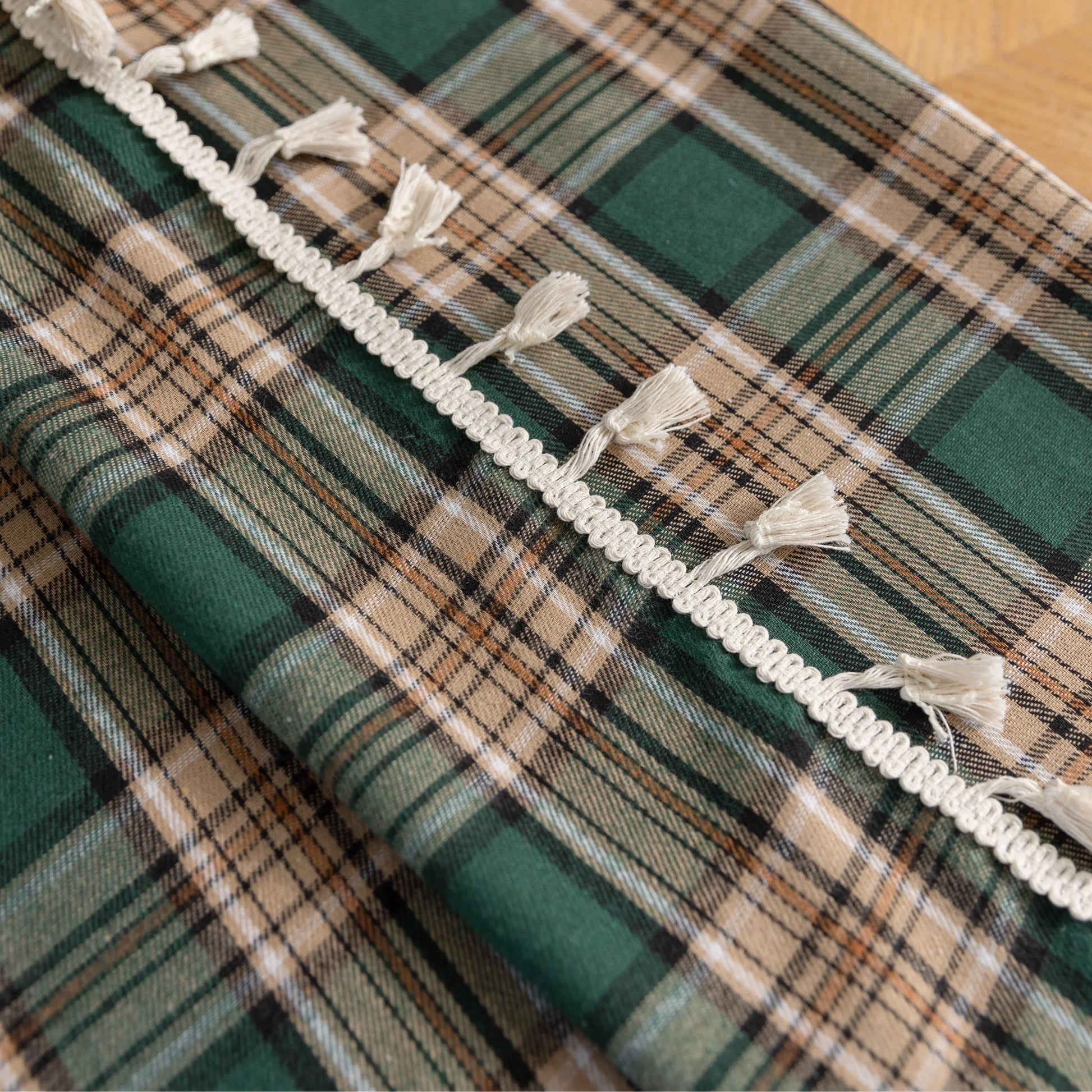 Green and beige plaid curtain fabric with a soft, woven texture, accented by decorative white tassel trim along the edge.
